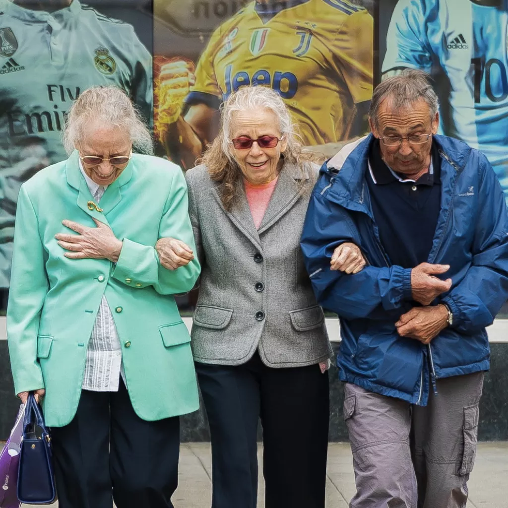 Three elderly people, arm in arm, crossing the road.